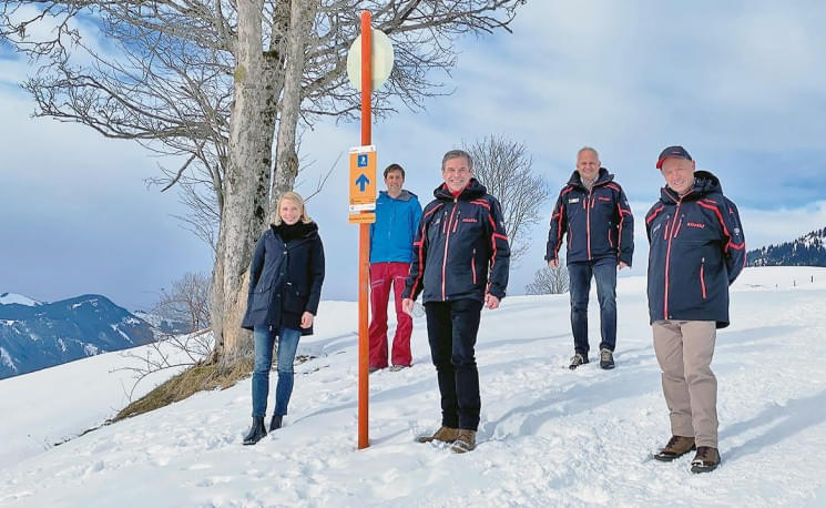 Fünf Personen stehen in Winterkleidung auf einem schneebedeckten Hügel mit Bäumen und einem Wegweiser. Der Himmel ist bewölkt, Berge im Hintergrund.