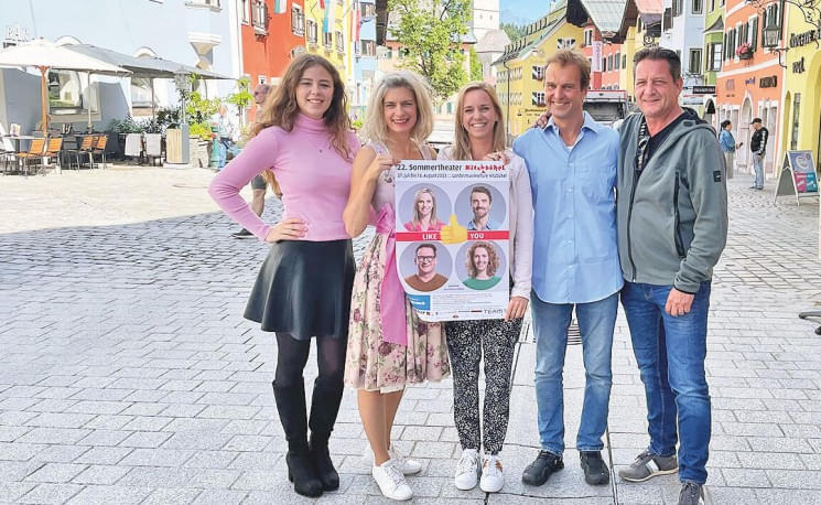 Fünf Personen stehen lächelnd auf einer Kopfsteinpflasterstraße, drei von ihnen halten ein Plakat mit Porträts und Text. Im Hintergrund sind bunte Gebäude sichtbar.