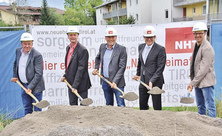 Fünf Männer in Bauhelmen halten Spaten bei einer Grundsteinlegung vor einem Werbebanner. Sie tragen Anzüge und stehen auf einer Baustelle mit Kieshaufen.