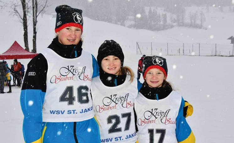 Drei junge Skifahrerinnen in bunter Ausrüstung stehen im Schnee, lächeln in die Kamera und tragen Rennwesten mit Nummern. Es schneit leicht im Hintergrund.