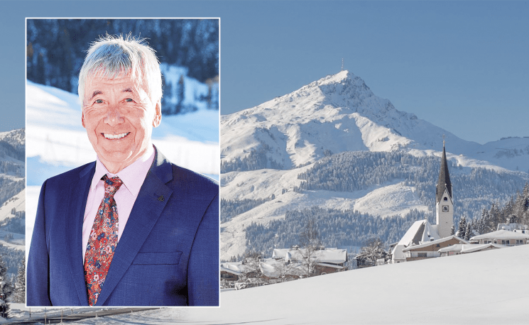 Links im Bild ein Mann im Anzug und Krawatte. Im Hintergrund eine verschneite Berglandschaft mit einem Kirchturm in einem Tal.