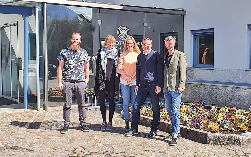 Fünf Personen stehen lächelnd vor einem Gebäude mit Glasfassade und Blumenbeet im Vordergrund. Sie tragen legere Kleidung und scheinen sich wohlzufühlen.