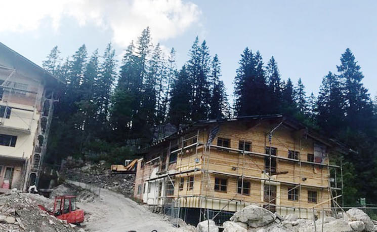 Rustikale Holzhütten im Bau vor einem Hintergrund von hohen Kiefern und blauem Himmel, umgeben von Bagger und Baumaterialien auf einer Bergbaustelle.