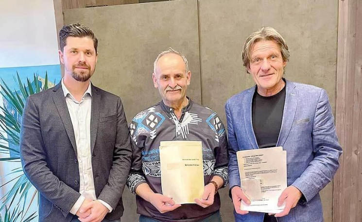 Drei Männer, zwei mit Zertifikaten in den Händen, stehen lächelnd nebeneinander in einem Raum mit Pflanzen und einfacher Einrichtung.