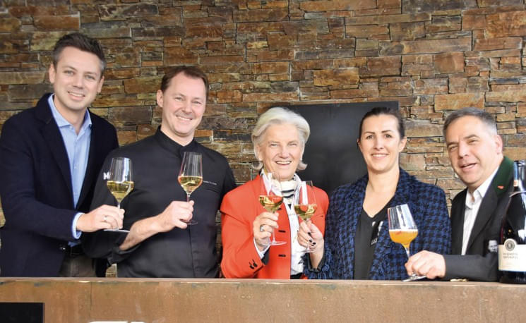 Fünf Personen in eleganter Kleidung stoßen mit Weingläsern an vor einer rustikalen Steinwand und lächeln in die Kamera.