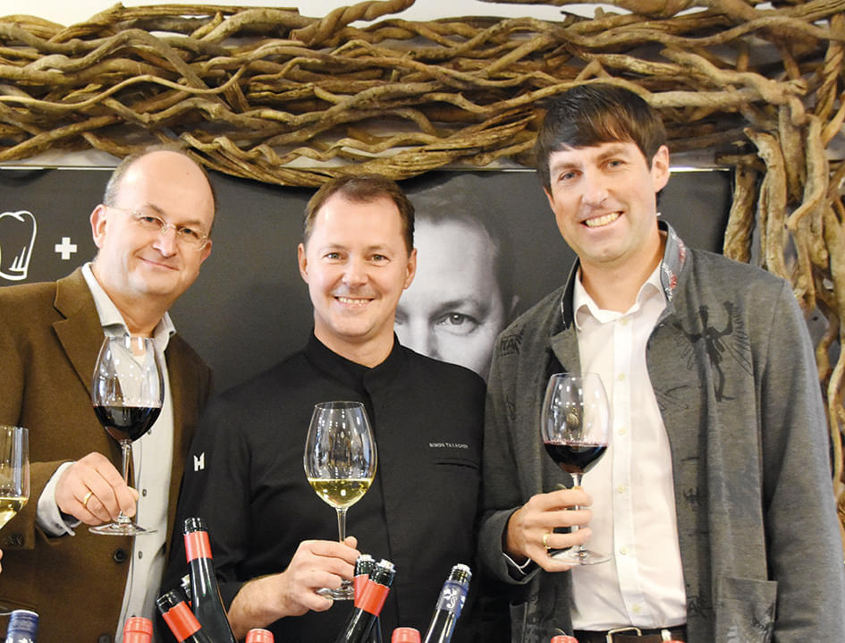 Drei Männer halten Weingläser vor einer dekorativen Wand mit Weinflaschen im Vordergrund, lächeln und stoßen an.