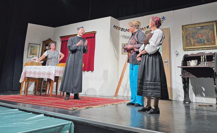 Eine Theatergruppe in Trachten führt eine humorvolle Szene auf, mit einem Tisch und Gemälden im Hintergrund. Alle Darsteller sind in traditionelle Kostüme gekleidet.