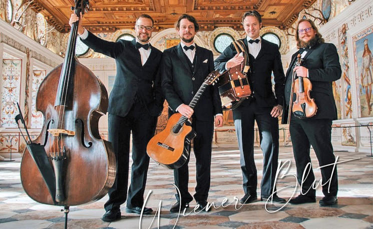 Vier Musiker in Anzügen posieren mit Instrumenten in einem eleganten, dekorativen Raum. Das Ambiente wirkt historisch und kunstvoll verziert.