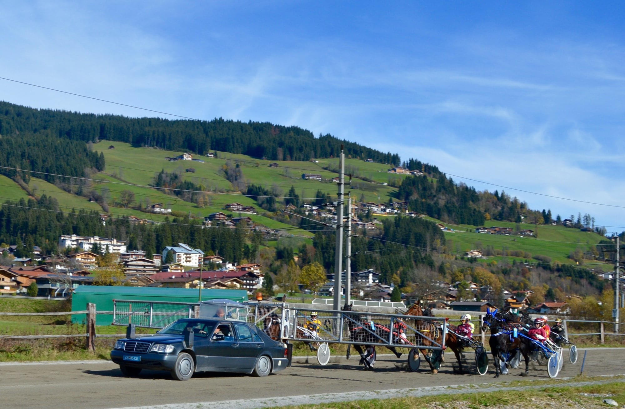 Pferdekutsche mit bunten Rädern fährt neben einem Auto durch eine ländliche Gegend mit grünen Hügeln und vereinzelten Häusern im Hintergrund.