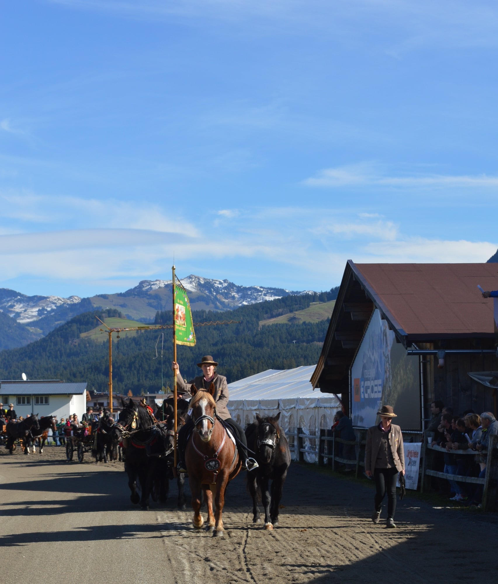 Ein Reiter in traditioneller Kleidung hält eine grüne Fahne auf einem Pferd. Im Hintergrund sind Alpen und ein Gebäude zu sehen, unter blauem Himmel.