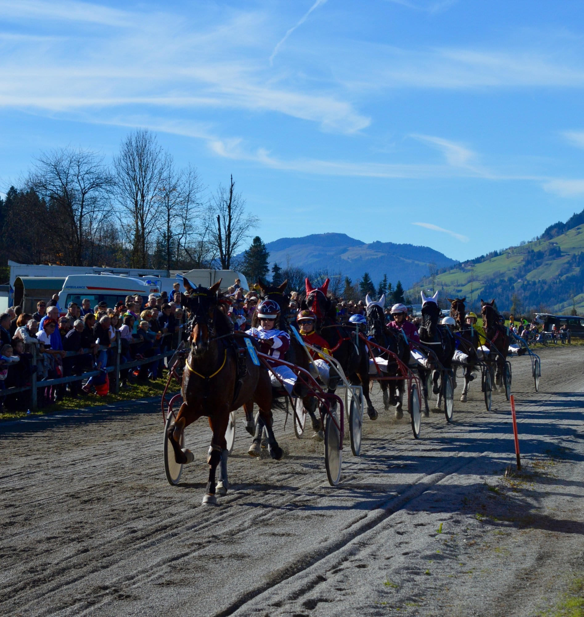 Mehrere Pferdekutschen rasen bei einem Rennen auf einer sandigen Strecke, umgeben von einer Menge Zuschauern und einer malerischen Berglandschaft im Hintergrund.