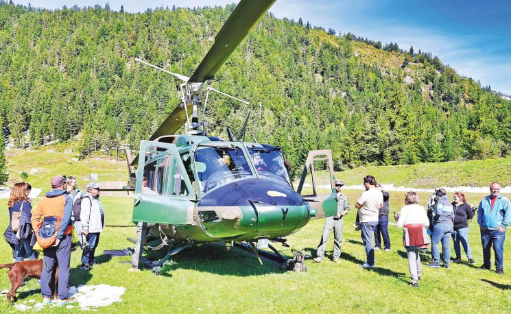 Ein grüner Militärhubschrauber steht auf einer Wiese in den Bergen. Menschen stehen um den Hubschrauber herum und betrachten ihn interessiert.