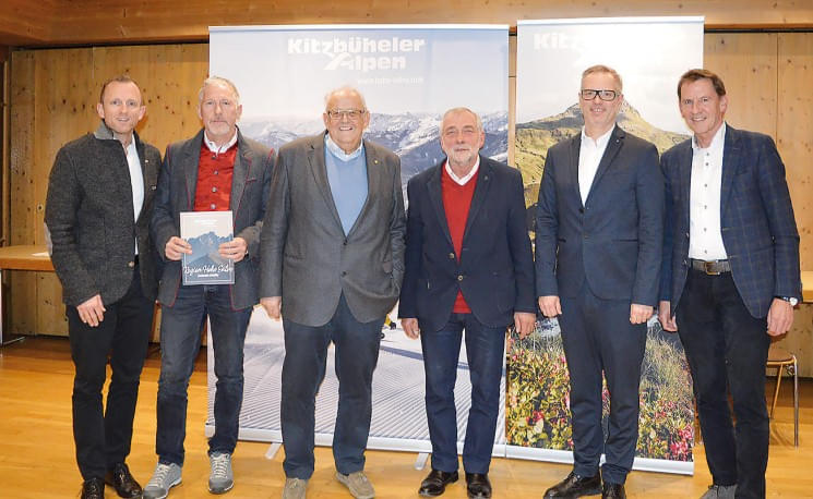 Sechs Männer in formeller Kleidung stehen lächelnd in einer Reihe vor Roll-up-Bannern in einem Raum mit Holzwänden.