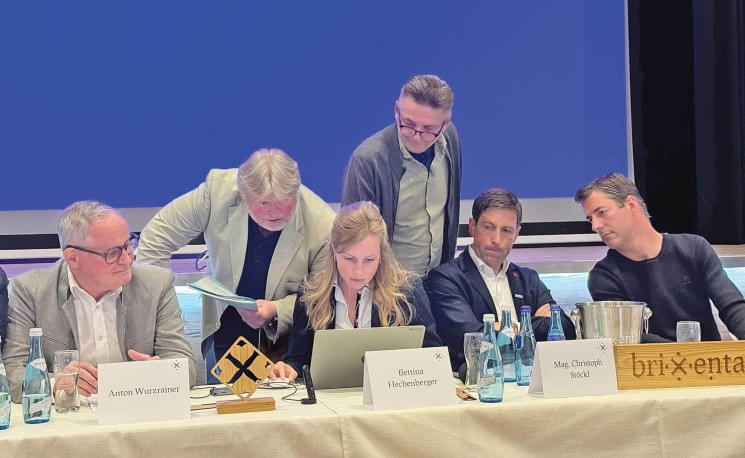 Sechs Personen sitzen an einem Tisch mit Getränken, während eine Frau auf einen Laptop schaut. Im Hintergrund steht ein Mann und zeigt auf den Bildschirm.