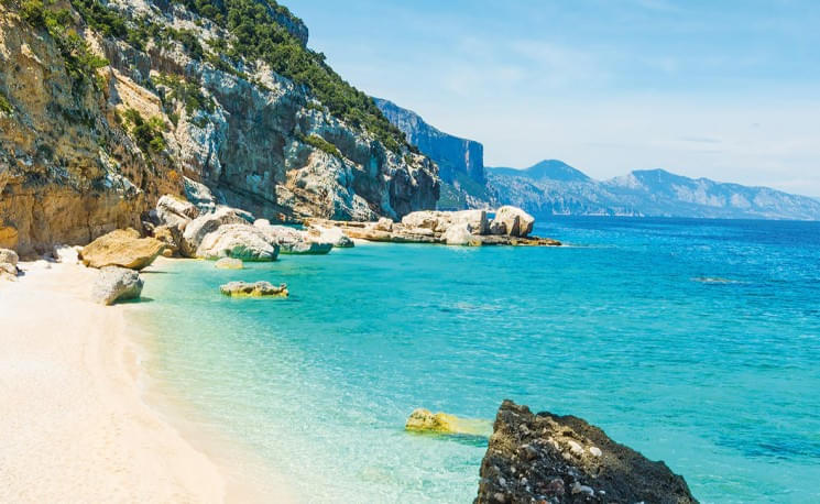 Wunderschöne Küstenlandschaft mit türkisfarbenem Wasser, weißem Sandstrand und beeindruckenden Felsen. Im Hintergrund erheben sich grüne Hügel und Berge.