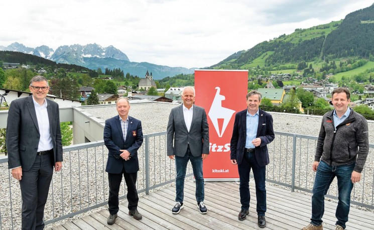 Gruppe von fünf Männern in Anzügen steht auf einer Terrasse mit alpiner Landschaft im Hintergrund, neben einem roten Banner mit der Aufschrift "KitzSki".