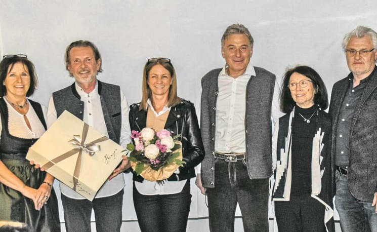 Sechs Personen stehen in einer Reihe, zwei halten Geschenke in den Händen, darunter Blumen und ein verpacktes Paket. Alle lächeln in die Kamera.