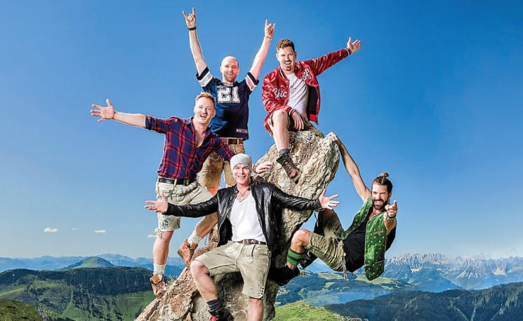 Fünf Männer in traditionellen bayerischen Trachten posieren fröhlich auf einem Felsen vor einer malerischen Bergkulisse bei klarem, sonnigem Wetter.