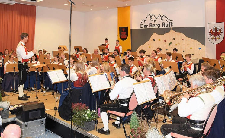 Eine Blaskapelle in traditioneller Tracht spielt auf einer Bühne. Im Hintergrund ist ein Plakat mit der Aufschrift "Der Berg Ruft" zu sehen.