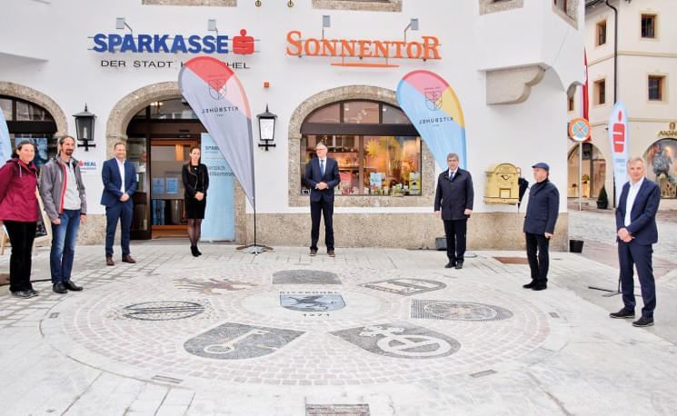 Eine Gruppe von Menschen steht vor einem Gebäude mit den Schildern 'Sparkasse' und 'Sonnentor', umgeben von zwei Fahnen und thematischen Pflastermarkierungen.