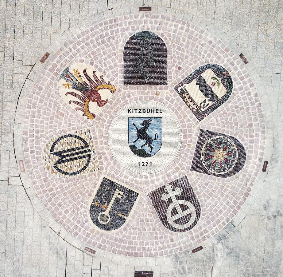 Ein rundes Mosaikpflaster zeigt Wappen und Motive. Zentral ein Schild mit "Kitzbühel" und Pferd, umgeben von Symbolen wie Schlüssel und Rad.