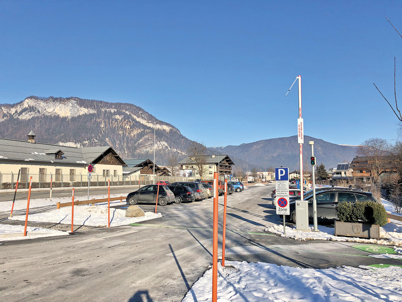 Winterliche Straßenszene in einer Bergregion mit Autos, schneebedecktem Boden und Häusern im Hintergrund, unter einem klaren blauen Himmel.