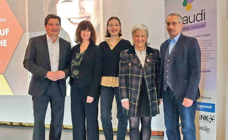 Fünf Personen stehen nebeneinander in einem Veranstaltungsraum und lächeln in die Kamera. Bei ihnen im Hintergrund sind ein Bildschirm und Logos zu sehen.