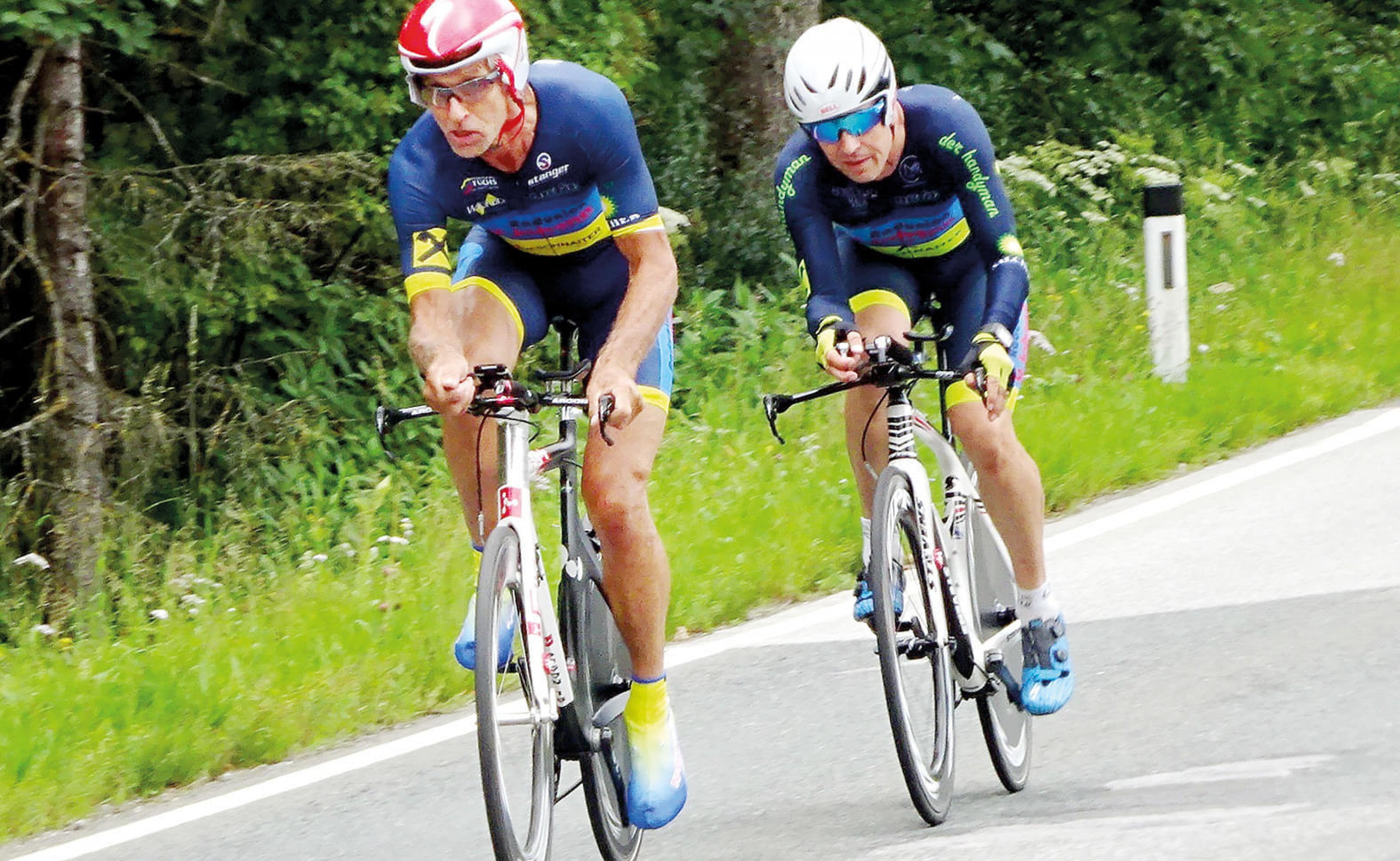 Zwei Radfahrer in Wettkampfausrüstung fahren schnell auf einer Landstraße durch eine grüne Landschaft, beide tragen Helme und Radtrikots mit blauen Details.