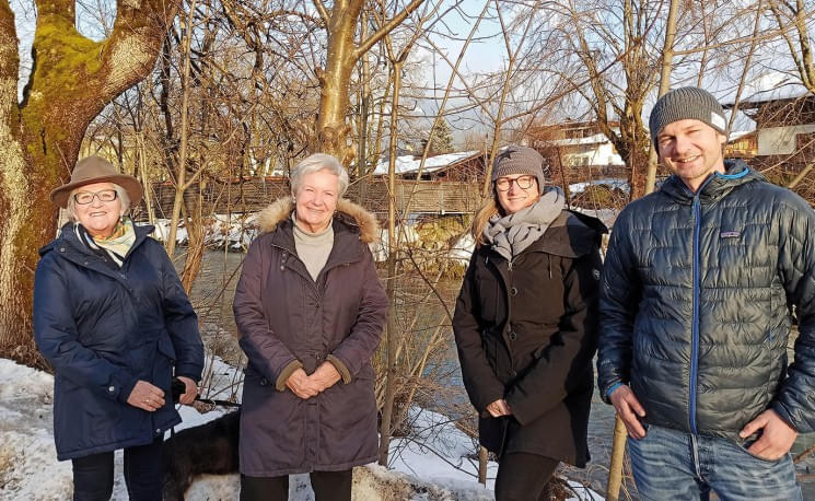 Vier Personen stehen lächelnd auf einem verschneiten Weg vor kahlen Bäumen. Sie sind warm gekleidet, es ist Winter. Im Hintergrund fließt ein Fluss.