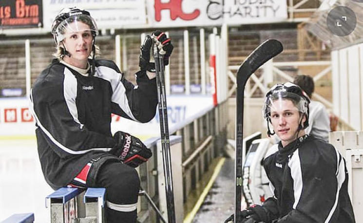 Zwei Eishockeyspieler in schwarzer Ausrüstung sitzen auf einer Bank am Spielfeldrand, halten ihre Schläger und tragen Helme mit Visier.