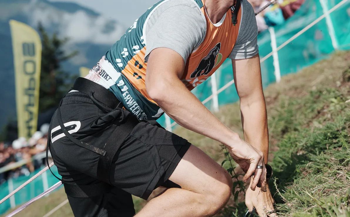 Ein Sportler in Action beim Berglauf, in leichter Vorwärtsneigung, wobei eine Hand den Boden berührt. Im Hintergrund sind Zuschauer und eine Fahne.