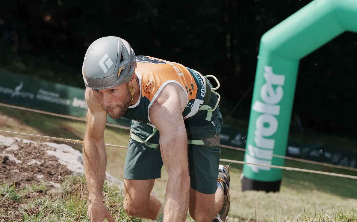Ein Sportler in Schutzhelm bewältigt einen Hindernisparcours unter freiem Himmel, während er einen Berg hinaufklettert. Im Hintergrund ist ein aufblasbares Banner.