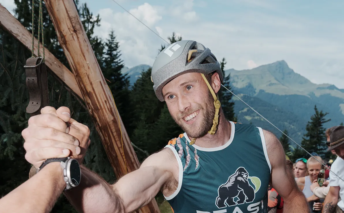 Ein Mann im Sport-Outfit mit Helm klatscht mit jemandem ab, während er auf einem Bergpfad läuft. Im Hintergrund sind Bäume und Berge zu sehen.