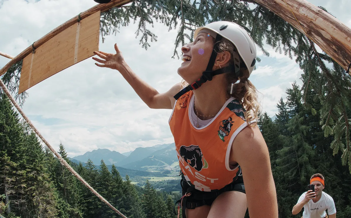Eine Person in sportlicher Ausrüstung und Helm klettert auf einem Baum mit Blick auf eine Berglandschaft. Eine andere Person im Hintergrund fotografiert.