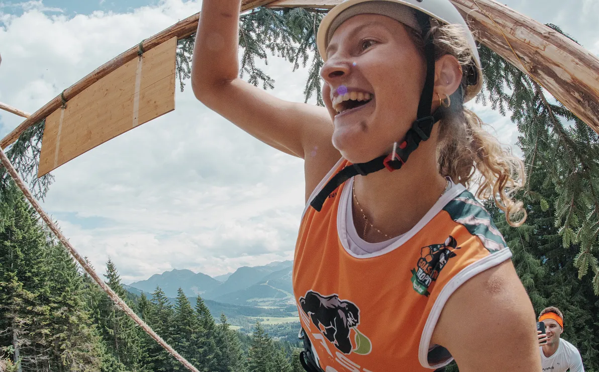 Frau beim Klettern in einem Hochseilgarten, lächelt und trägt Helm. Im Hintergrund sind Bäume und Berge unter blauem Himmel sichtbar.