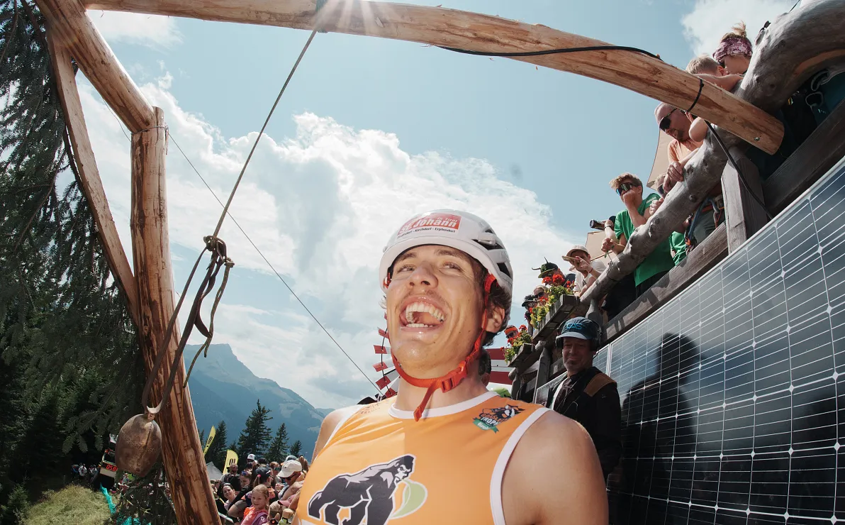 Ein Mann in Sportkleidung und Helm lacht breit bei einem Outdoor-Event in den Bergen. Zuschauer sind auf einer Tribüne im Hintergrund zu sehen.
