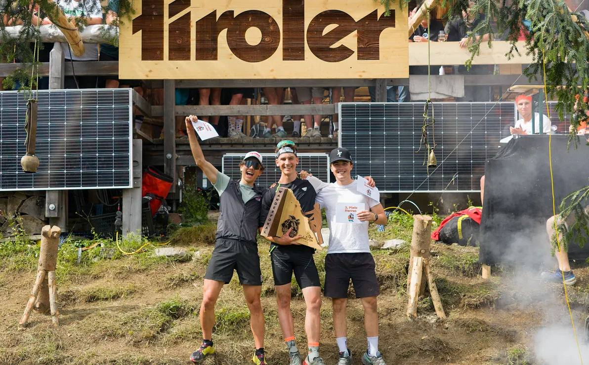 Drei fröhliche Personen posieren vor einem Gebäude mit "tiroler" Schild, umgeben von Solarpaneelen und grüner Natur, möglicherweise bei einem Sportevent.