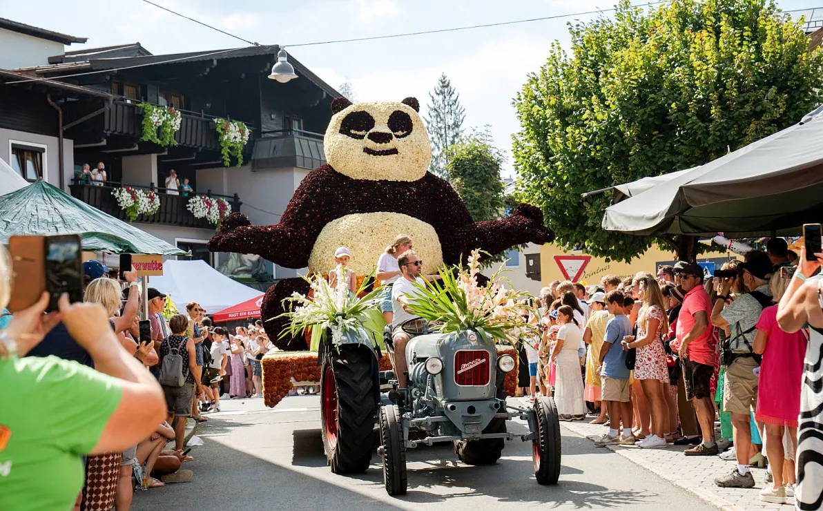 Ein Festzug mit einem Traktor, der einen großen Panda aus Blumen zieht, umgeben von Menschenmengen bei sonnigem Wetter auf einer Straße.