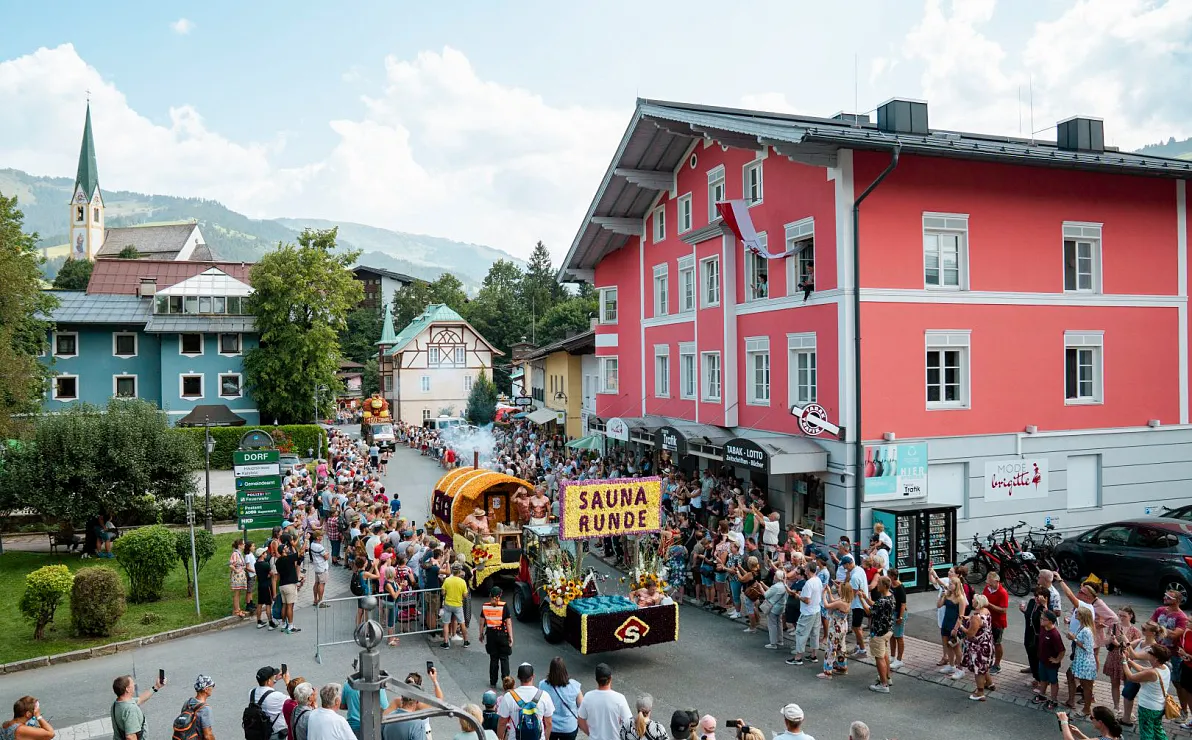Eine fröhliche Straßenparade in einer malerischen Stadt mit bunten Gebäuden, vielen Zuschauern und einem Umzugswagen mit dem Banner "Sauna Runde".
