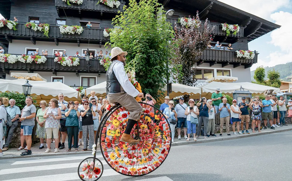 Ein Mann in historischer Kleidung fährt ein großes Hochrad, verziert mit Blumen, vor einem Publikum und traditionellen Häusern mit Blumenschmuck.