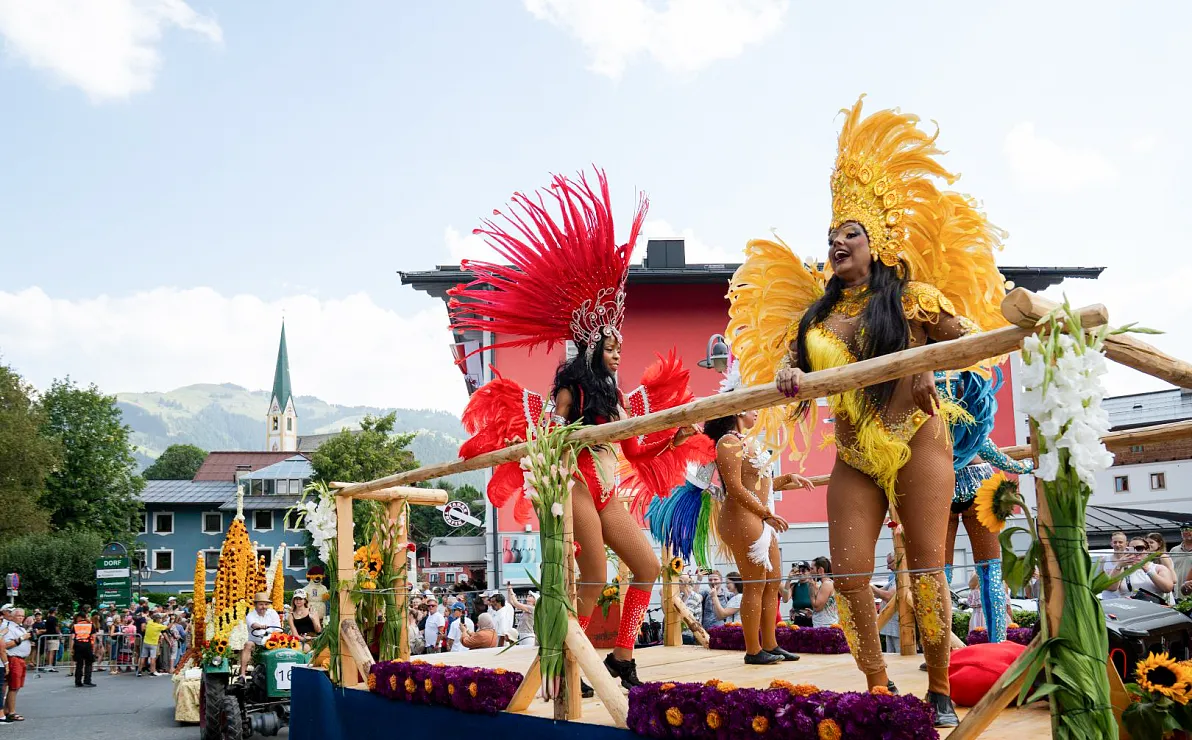 Farbenfrohe Szene auf einem Umzugswagen mit Tänzerinnen in bunten Kostümen mit Federschmuck. Hintergrund zeigt eine Kirche und Berge.
