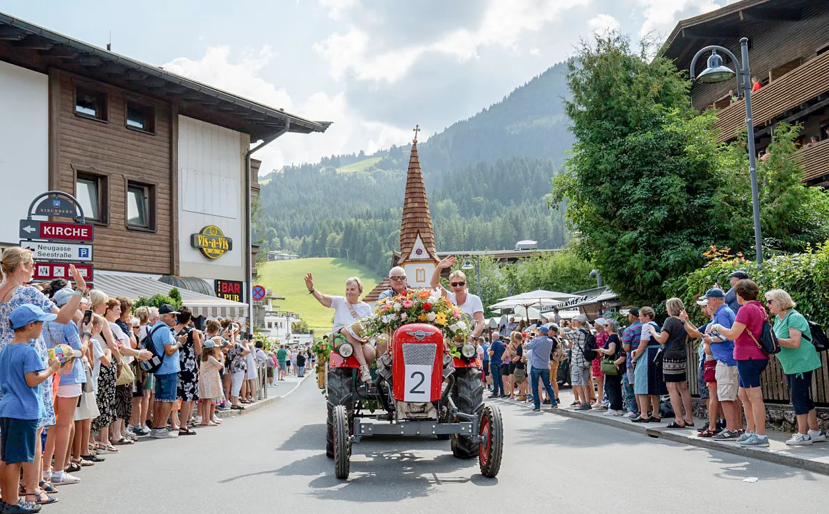 Festlich geschmückter Traktor mit Blumen fährt durch eine mit Menschen gesäumte Straße in einer alpinen Umgebung bei sonnigem Wetter.