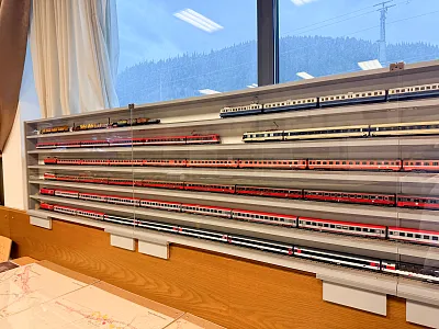 Ein Regal voller maßstabsgetreuer Modellzüge in verschiedenen Farben und Stilen steht vor einem Fenster mit Blick auf eine schneebedeckte Landschaft.