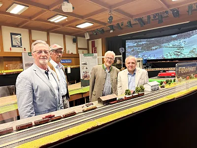 Vier Männer stehen in einer Ausstellungshalle neben einem Modellbahn-Layout mit Zügen und Landschaften. Im Hintergrund sind weitere Modelle und Poster sichtbar.
