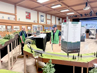 Zwei Männer in grünen Westen stehen neben einer Modelleisenbahn-Anlage in einem Ausstellungsraum mit hölzerner Decke und Bildern an den Wänden.