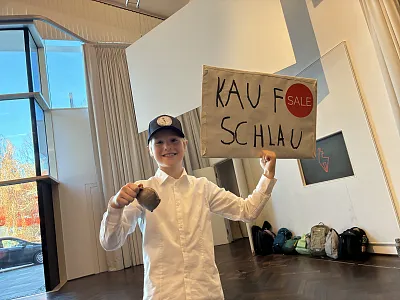 Ein Junge in weißem Hemd und Baseballkappe hält ein Schild mit der Aufschrift "Kauf schlau" und einem roten Sale-Aufkleber in einem hellen Raum.