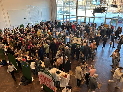 Menschenmenge auf einer Messe in einer großen Halle mit Informationsständen und bunten Plakaten. Besucher sprechen miteinander und betrachten die Ausstellungen.