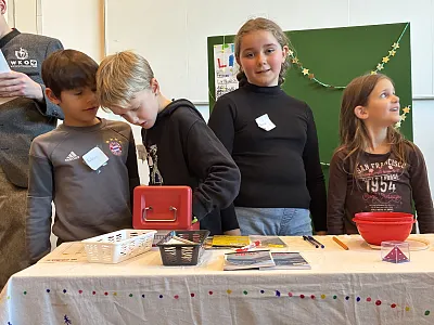 Vier Kinder stehen hinter einem Tisch mit Bastelmaterialien und lächeln. Im Hintergrund sind eine Tafel und ein Poster zu sehen.