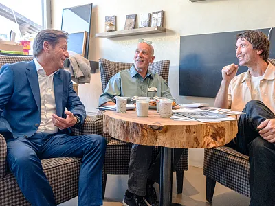 Drei Männer sitzen fröhlich an einem runden Holztisch in einem Café und unterhalten sich. Sie halten Tassen in der Hand und wirken entspannt.