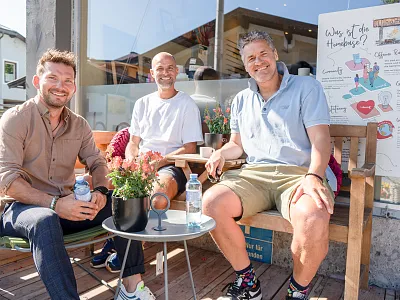 Drei Männer sitzen lächelnd auf einer Holzbank vor einem Café. Sie tragen Freizeitkleidung, und auf dem Tisch stehen Pflanzen und Wasserflaschen.